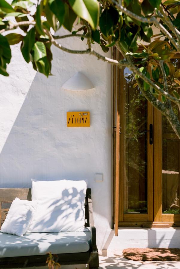 10 AUTHENTIC Finca Hotels in Ibiza 23 A cozy outdoor bench with white cushions sits near a wooden door on a whitewashed wall, shaded by leafy branches—evoking the relaxed charm of finca hotels in Ibiza. Sunlight casts shadows, and a yellow house number sign is mounted above the bench.
