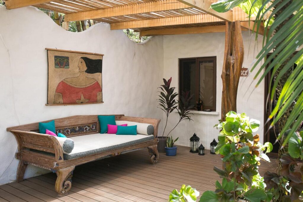 Unique Hotels in Costa Rica: Top Stays 9 A cozy outdoor patio at one of the unique hotels in Costa Rica features a wooden daybed with colorful pillows, potted plants, a white stucco wall, a window, hanging lanterns, and a painting of a woman under a wooden pergola.