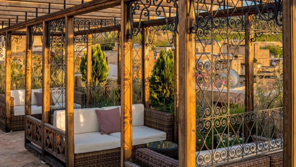 7 UNIQUE Riads in Fez You Won't Forget 10 Outdoor seating area with wicker sofas and white cushions under a wooden pergola with decorative wrought iron panels, reminiscent of the charming riads in Fez. Green plants and a rustic stone background are visible in warm sunlight.