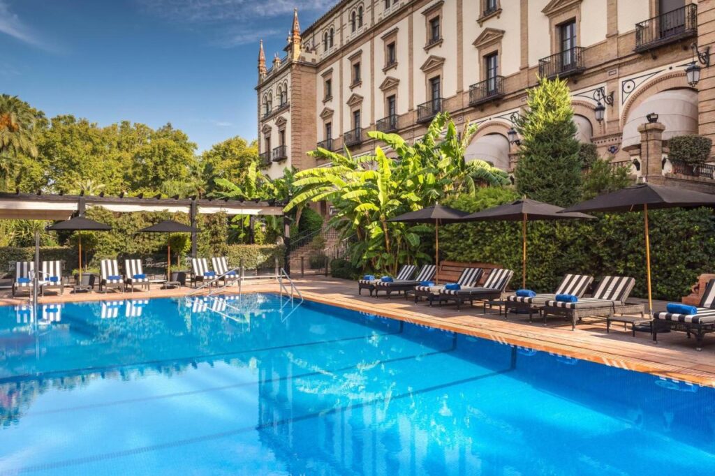 10 UNIQUE Hotels with Pool in Seville 22 A luxurious outdoor swimming pool at one of the top hotels with pool in Seville, featuring striped lounge chairs and black umbrellas, surrounded by lush greenery and a grand, historic building with balconies under a blue sky.