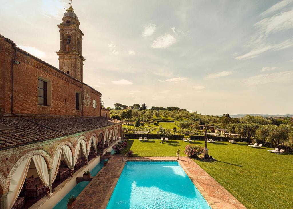7 Best Agriturismos in Tuscany 12 A serene outdoor pool beside a historic brick building with arches and a bell tower, overlooking a lush green lawn and rolling hills—a setting reminiscent of the best agriturismos in Tuscany, all beneath a partly cloudy sky.
