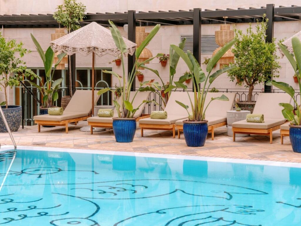 10 UNIQUE Hotels with Pool in Seville 27 Four lounge chairs with beige cushions and green towels sit beside a clear blue pool, surrounded by large potted plants and an umbrella, evoking the tranquil ambiance found at top hotels with pool in Seville.