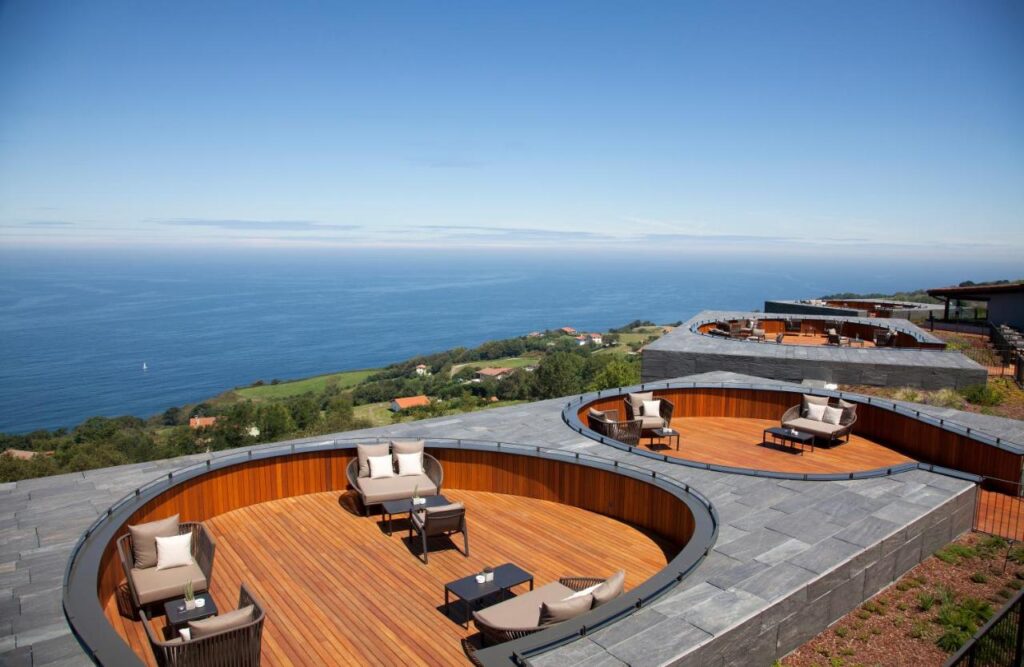Best Boutique Hotels in San Sebastian 27 Circular wooden terraces with outdoor seating overlook a scenic coastline and ocean under a clear blue sky—an inviting scene often found at boutique hotels in San Sebastian, with green hills and a few buildings visible below.