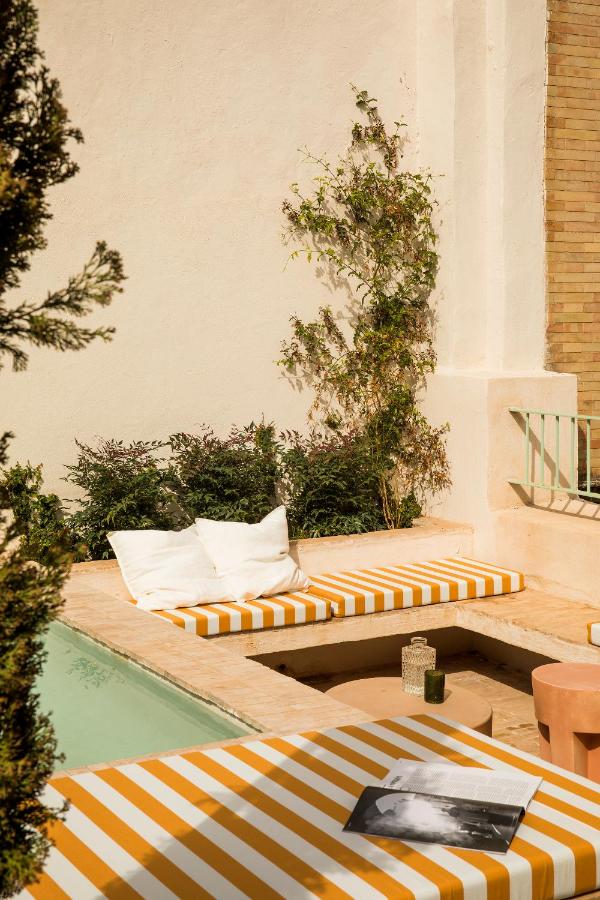 10 UNIQUE Hotels with Pool in Seville 38 A cozy outdoor patio at one of the charming hotels with pool in Seville, featuring yellow and white striped cushions on lounge beds beside a small pool, white pillows, a book, a candle, and greenery against cream and brick walls.