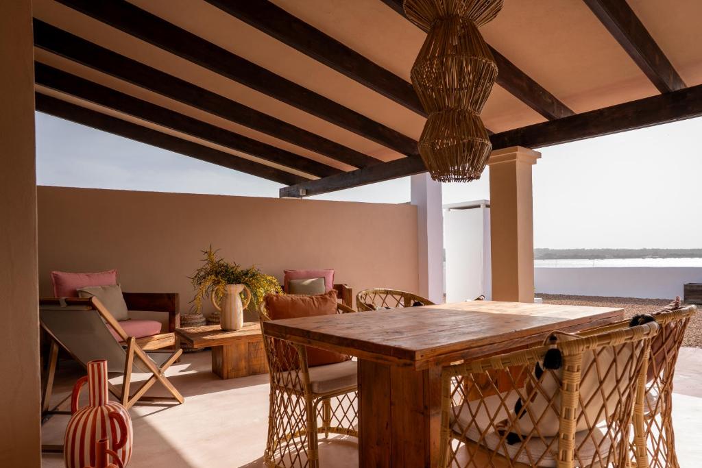 A covered outdoor patio reminiscent of boutique hotels in Formentera features wooden beams, wicker chairs around a wooden table, and cozy cushioned seating. Decorative vases and woven light fixtures create a warm atmosphere with water views in the background.