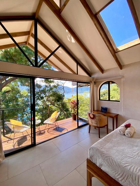 8 UNIQUE Places Where to Stay in Lake Atitlán 28 A bright, airy bedroom with exposed beams and large glass doors opens to a balcony overlooking trees and a lake under a blue sky—an ideal spot if you’re searching for where to stay in Lake Atitlán. Inside, there’s a cozy bed and a desk with a chair.