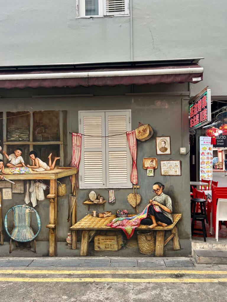 13 Most Unique Hotels in Singapore for an Incredible Holiday 58 A street mural near unique hotels in Singapore shows a woman sewing at a wooden table while children peek from a window, all painted on a gray wall beside a restaurant with red chairs and a Chinese sign.
