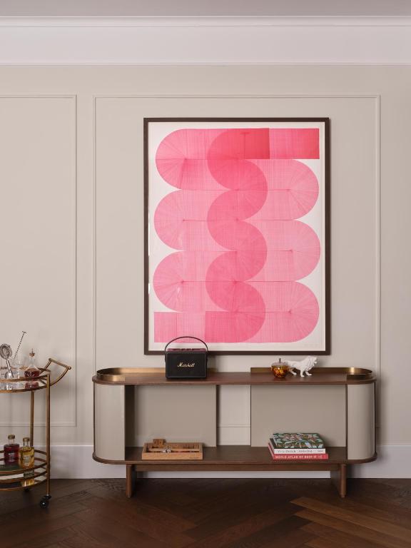 Authentic Boutique Hotels in Munich 31 A framed abstract artwork with overlapping pink shapes hangs above a console table, echoing the chic vibe of boutique hotels in Munich. On the table are a black radio, white decorative object, books, and a small bar cart with bottles to the side.