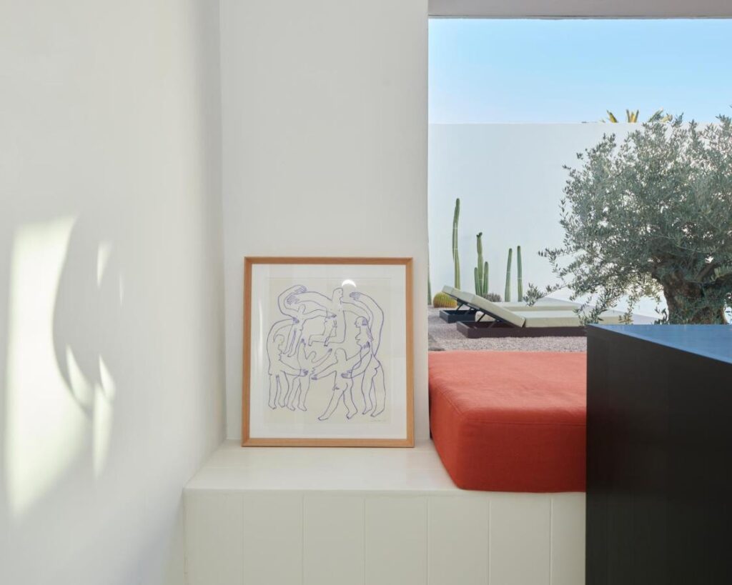 Adults Only Hotels in Fuerteventura 5 A minimalist room in the style of adults only hotels in Fuerteventura, with a framed abstract line drawing on a white ledge, a red cushion bench, and a large window overlooking plants and a lounge chair outside.