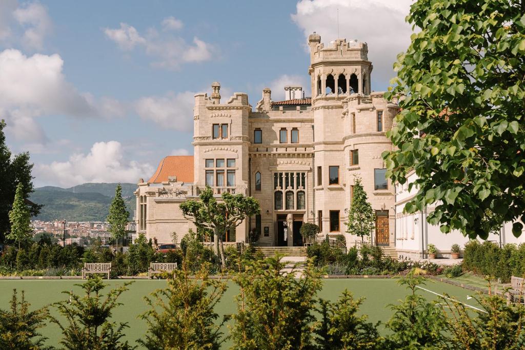 8 Unique Boutique Hotels in Bilbao Spain 22 A grand, historic stone mansion with towers and ornate windows stands amid lush greenery, reminiscent of the boutique hotels in Bilbao, with mountains and a city visible in the background under a partly cloudy sky.