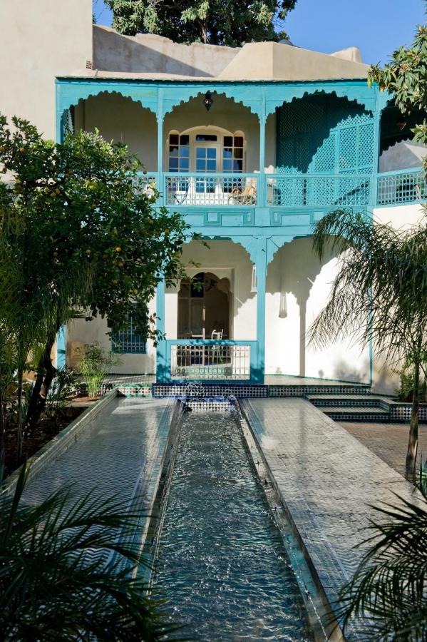 7 UNIQUE Riads in Fez You Won't Forget 11 A narrow swimming pool runs through a lush garden toward a two-story building with turquoise wooden balconies and intricate arches, capturing the tranquil and inviting courtyard scene typical of traditional riads in Fez.