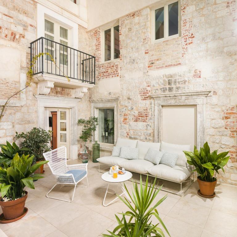 8 TOP Boutique Hotels in Split 6 A cozy outdoor patio with white stone walls, a white sofa, a white chair, and potted green plants—perfect for relaxing in Split. Large windows and a small balcony overlook this inviting seating area, an ideal option when considering where to stay in Split.