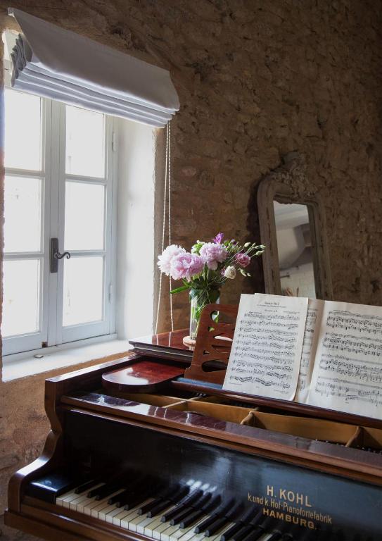 11 Unique Places to Stay in Provence 45 A vase of pink flowers and an open music book sit on a piano near a sunlit window, with a rustic stone wall and mirror in the background—capturing the charm of unique places to stay in Provence.