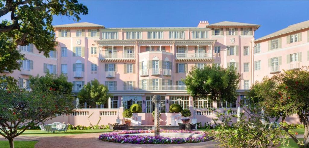 Unique Places to Stay in Cape Town 6 A historic four-story pink hotel with white trim stands behind a courtyard garden featuring a circular flower bed, stone fountain, trees, and patio seating—one of the most unique places to stay in Cape Town under a clear blue sky.