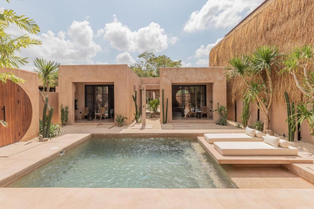 A modern outdoor pool, like those at the best family villas in Bali, is framed by beige stucco buildings, cacti, palm trees, and two white loungers beneath a bright blue sky with scattered clouds.