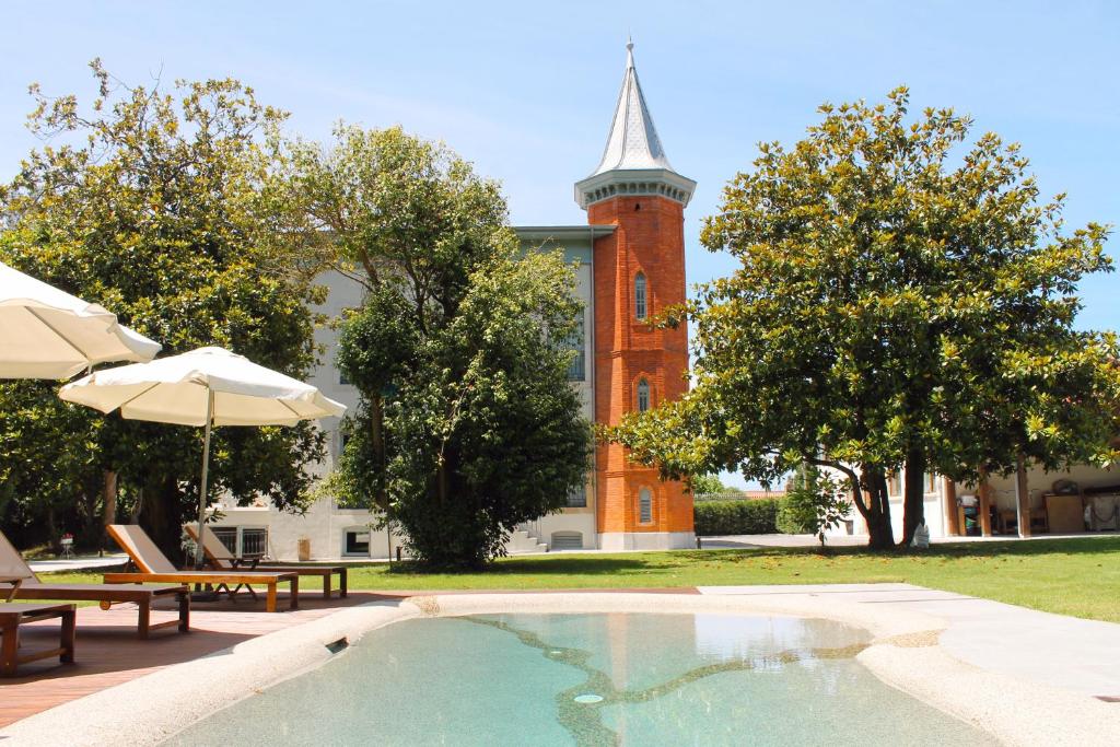 A small, clear swimming pool sits in a sunny yard with lounge chairs and umbrellas, capturing the charm of boutique hotels in Asturias. In the background, a tall red-brick tower rises among lush green trees surrounding the area.