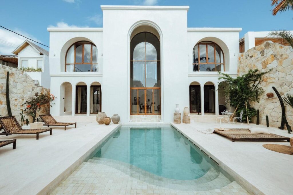 14 UNIQUE Boutique Hotels in Canggu, Bali 14 Modern white villa with arched windows and doors, featuring a central swimming pool surrounded by lounge chairs, stone walls, and tropical plants—perfectly capturing the stylish charm of boutique hotels in Canggu under a blue sky.