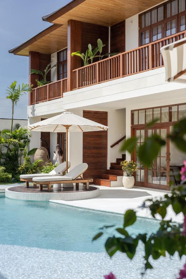 14 UNIQUE Boutique Hotels in Canggu, Bali 41 A modern villa with large wooden windows and balconies overlooks a clear blue swimming pool, reminiscent of boutique hotels in Canggu. Two lounge chairs with an umbrella sit by the pool, lush plants decorate the area, and a person stands on the balcony.