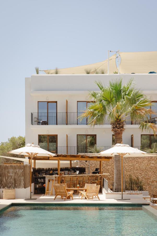 A modern white building with balconies overlooks a pool surrounded by palm trees, sun umbrellas, and outdoor wooden furniture—an inviting scene typical of boutique hotels in Formentera on a sunny day.