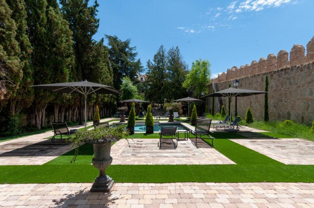 25 Weekend Trips from Madrid Max. 2h Away 99 A paved patio with lounge chairs and umbrellas surrounds a small pool—perfect for relaxing on weekend trips from Madrid. Tall trees and a stone wall enclose the area, while patches of green artificial grass add color to the courtyard.