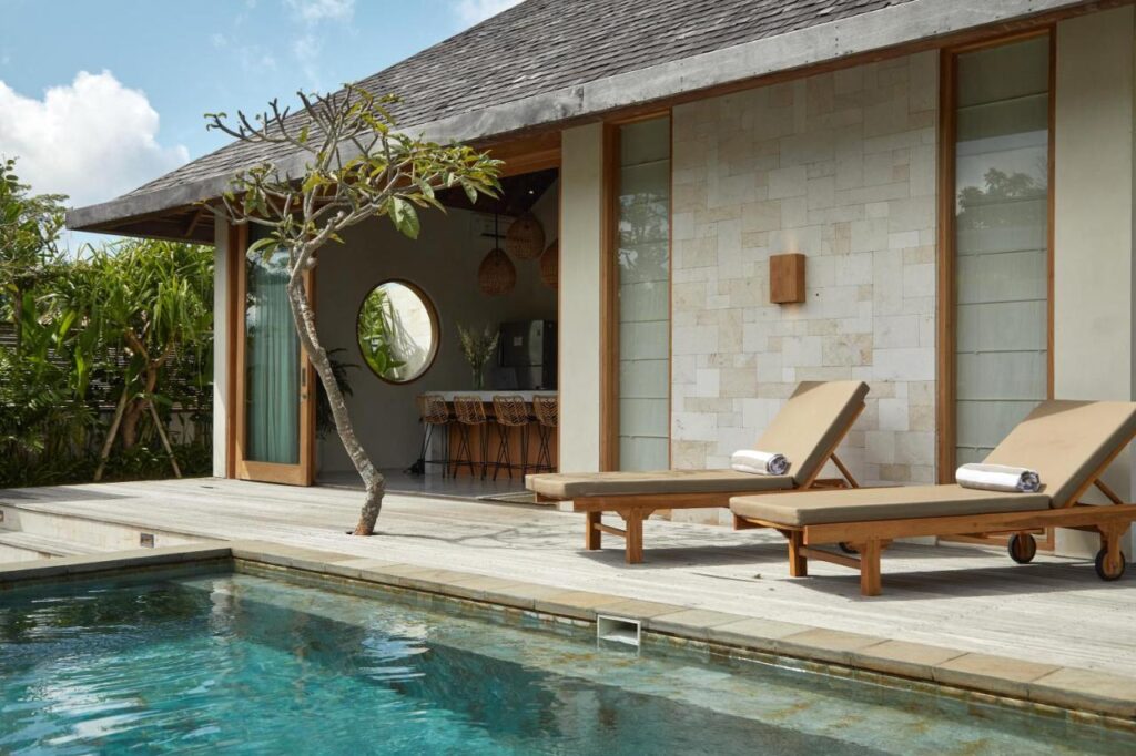 Modern villa with stone walls, wooden deck, and two beige lounge chairs beside a clear swimming pool. Sliding glass doors open to an airy interior with a round window and bar stools—one of the best family villas in Bali, surrounded by tropical plants.
