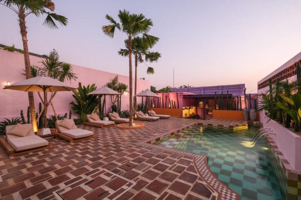 14 UNIQUE Boutique Hotels in Canggu, Bali 48 A stylish outdoor pool area at sunset with palm trees, white lounge chairs and umbrellas, patterned tile flooring, and pastel pink and purple walls—reminiscent of the chic designs found in boutique hotels in Canggu.
