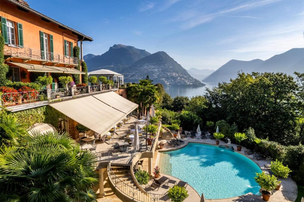 A luxurious villa with a large pool overlooks a serene lake surrounded by mountains—much like the best hotels in Lugano. Terraces with tables, umbrellas, and lush greenery create a relaxing, scenic atmosphere under a clear blue sky.