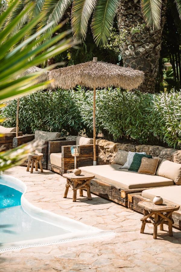 10 AUTHENTIC Finca Hotels in Ibiza 7 A sunlit poolside lounge area at one of the charming finca hotels in Ibiza, with rustic wooden sofas, beige cushions, small wooden stools, and straw umbrellas, surrounded by lush green plants and palm trees.