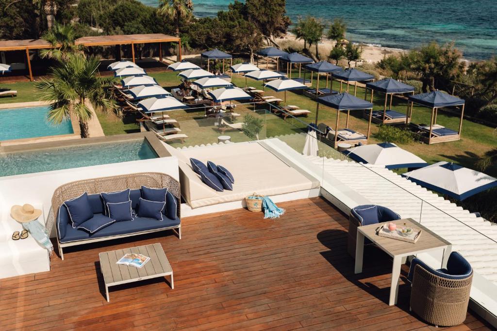 A stylish outdoor lounge area with blue and white furniture overlooks a pool, rows of sunbeds with umbrellas, grassy lawns, and the sea beyond—evoking the chic ambiance of boutique hotels in Formentera.