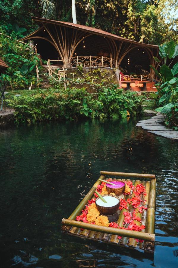 13 Most UNIQUE Places to Stay in Ubud 11 A bamboo raft with plates of colorful food floats on a pond surrounded by lush greenery. In the background, a wooden, open-air dining area with curved roofing overlooks the water—one of the unique places to stay in Ubud.