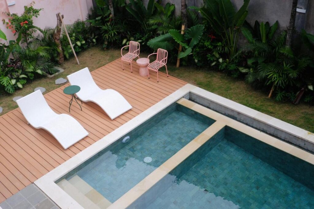 14 UNIQUE Boutique Hotels in Canggu, Bali 31 A modern backyard reminiscent of boutique hotels in Canggu, featuring a rectangular swimming pool, two white lounge chairs, a small green table, two pink chairs, and lush green plants surrounding the deck area.