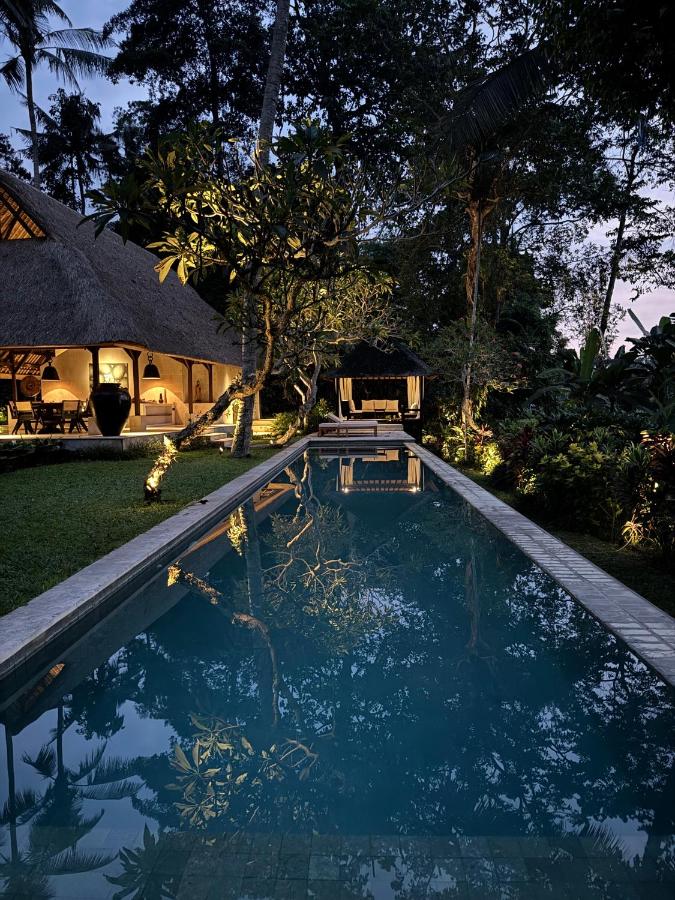 A tranquil pool stretches into a lush garden at dusk, reflecting trees and soft lights from a thatched-roof villa—one of the best family villas in Bali—surrounded by tropical plants, an open lounge area, and dense greenery.