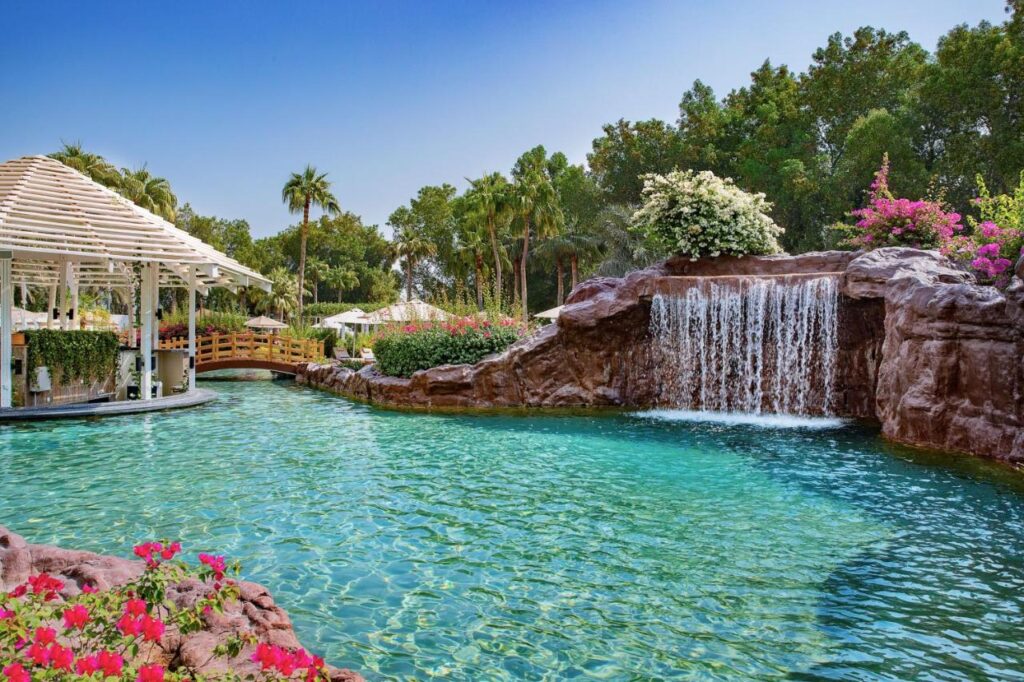 Where to Stay in Doha, Qatar: Transit, City & Beach Hotels 16 A resort pool, ideal for those seeking where to stay in Doha, features a waterfall flowing over rocks, lush greenery, palm trees, and vibrant flowers. A gazebo sits to the left, partly over the water under a clear blue sky.
