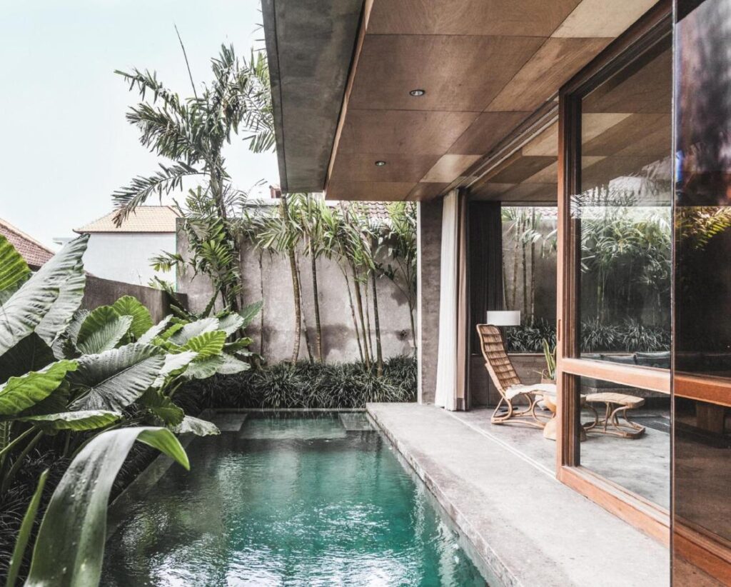 14 UNIQUE Boutique Hotels in Canggu, Bali 23 Modern villa with a private pool, lush tropical plants, a concrete patio, and floor-to-ceiling glass doors—offering the tranquil, resort-like vibe of boutique hotels in Canggu. Wooden furniture adds to the serene atmosphere inside.