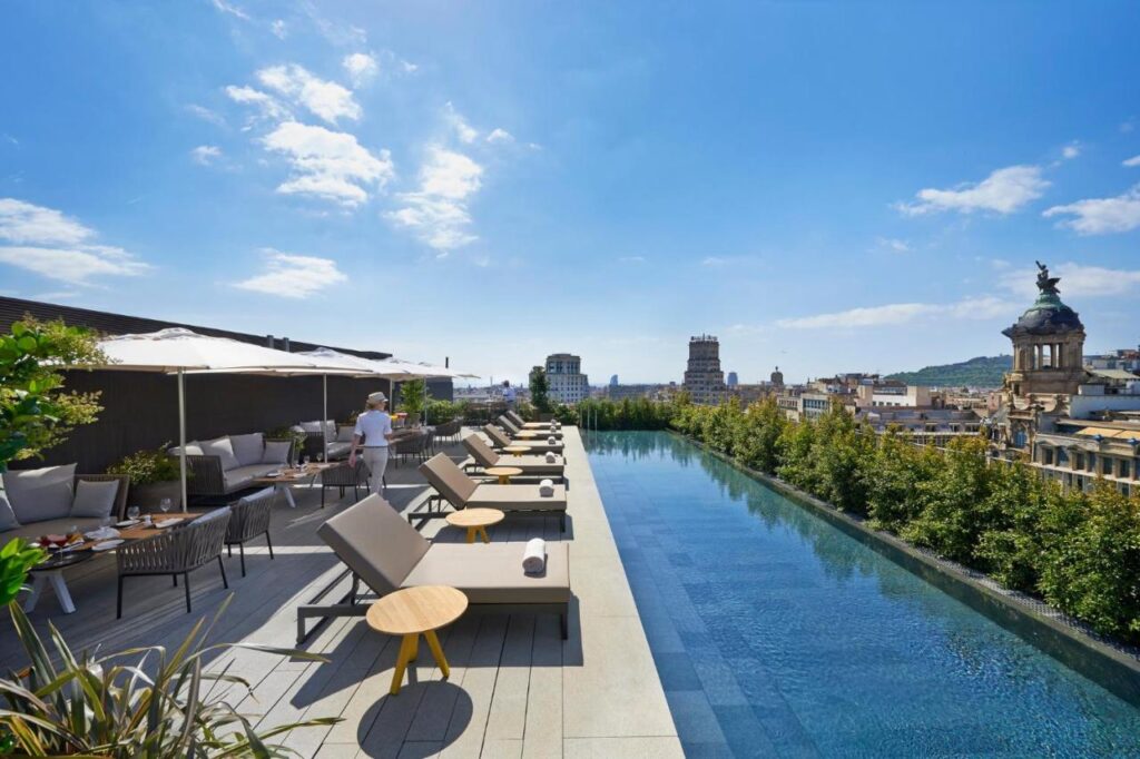 Top Barcelona Hotels with Rooftop Pool 20 A rooftop pool with lounge chairs and umbrellas overlooks a cityscape under a bright blue sky. A person stands near the pool, surrounded by greenery and historic buildings—a stunning scene at one of the top Barcelona Hotels with Rooftop Pool.