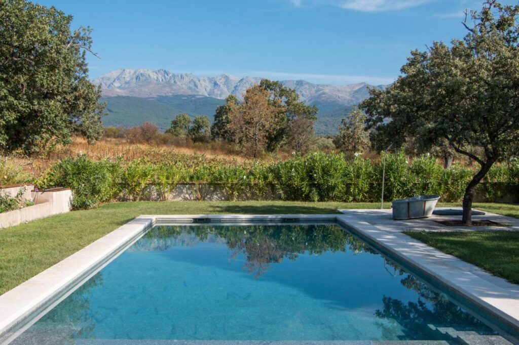 25 Weekend Trips from Madrid Max. 2h Away 5 A clear swimming pool with a stone border sits in a grassy yard, surrounded by trees and bushes, perfect for relaxing on weekend trips from Madrid, with mountains and a blue sky in the background.