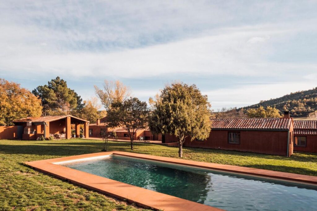 25 Weekend Trips from Madrid Max. 2h Away 49 A rectangular outdoor swimming pool sits on a grassy lawn in front of a rustic, terracotta-colored house—an idyllic spot for weekend trips from Madrid—surrounded by trees and hills under a partly cloudy sky.