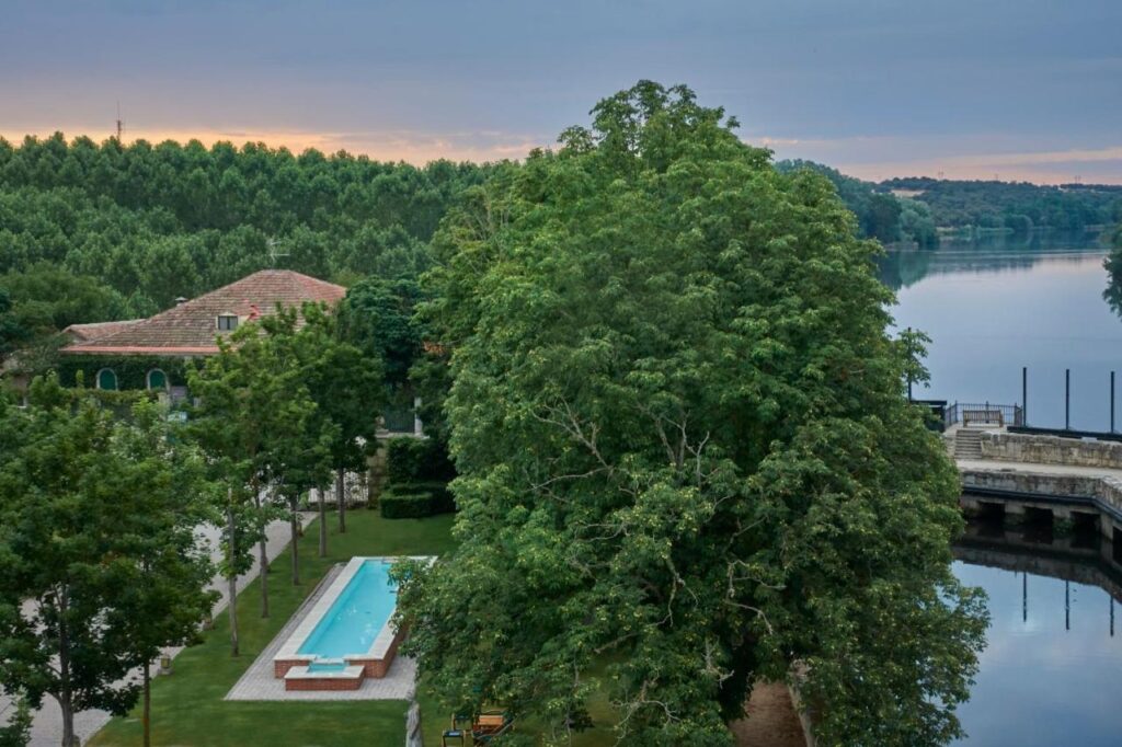 25 Weekend Trips from Madrid Max. 2h Away 25 A rectangular swimming pool is surrounded by green trees and a grassy area near a river, perfect for relaxing weekend trips from Madrid, with a red-tiled roof building nearby and a wooded landscape in the background under a cloudy sky.