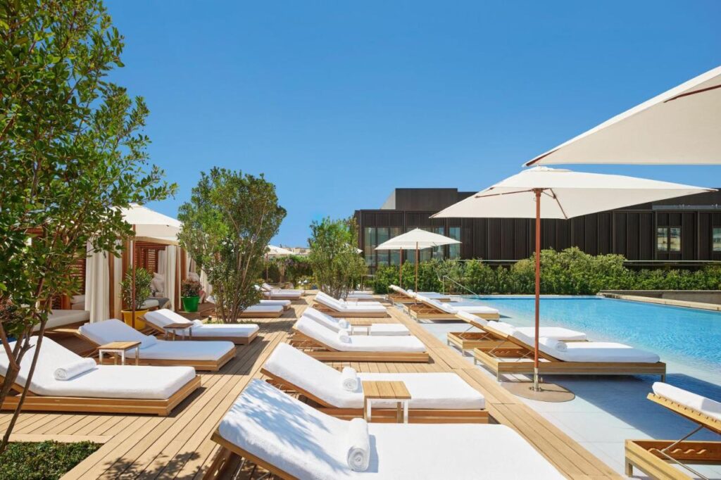 Where to Stay in Madrid: 14 Best Hotels 20 A row of white cushioned lounge chairs and umbrellas lines a wooden deck beside a clear blue swimming pool—an inviting spot reminiscent of the luxury escapes you'll find when searching where to stay in Madrid first time, surrounded by greenery under a bright sky.
