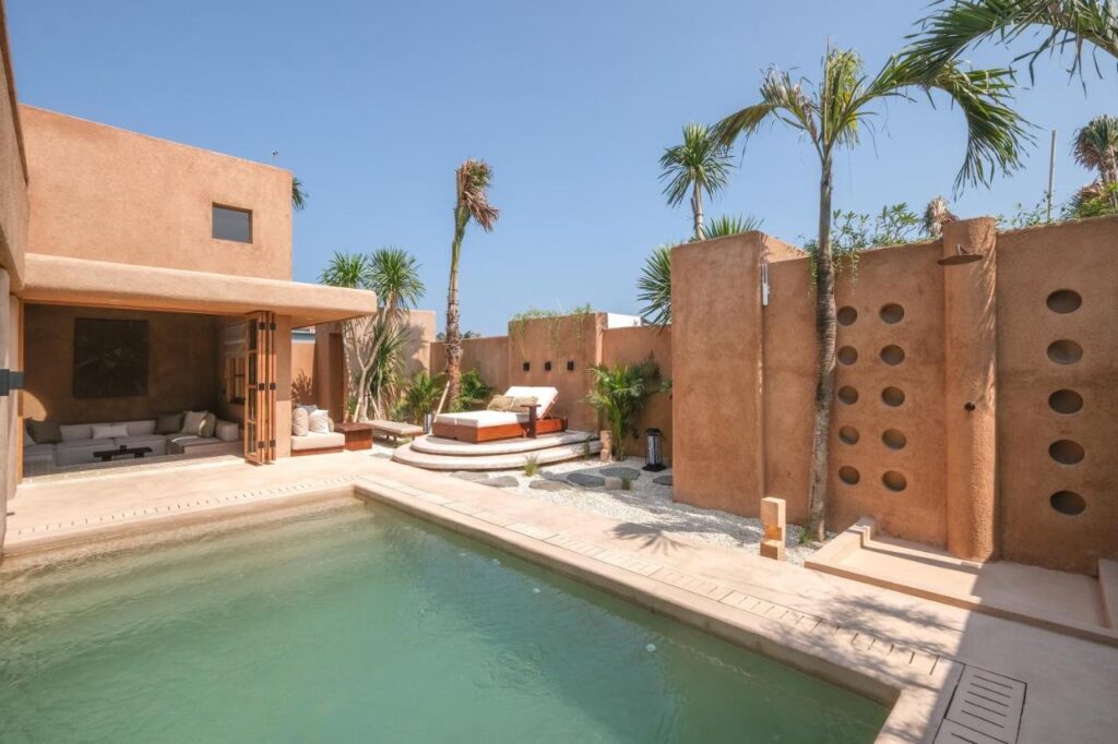 A modern villa with tan stucco walls, often ranked among the best family villas in Bali, features a private pool, lounge chairs, palm trees, and an outdoor seating area under an open sky. Large round cutouts decorate one of the walls.