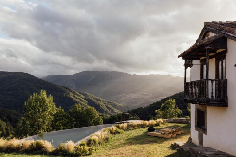 A rustic house with a wooden balcony, like those found in boutique hotels in Asturias, overlooks a serene infinity pool, lounge chairs, and lush green mountains under a cloudy sky at sunset.