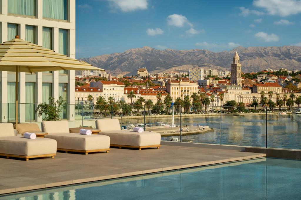 8 TOP Boutique Hotels in Split 20 A rooftop pool with sun loungers overlooks a coastal city with historic buildings—an ideal spot for those wondering where to stay in Split, surrounded by palm trees and mountains under a clear blue sky.