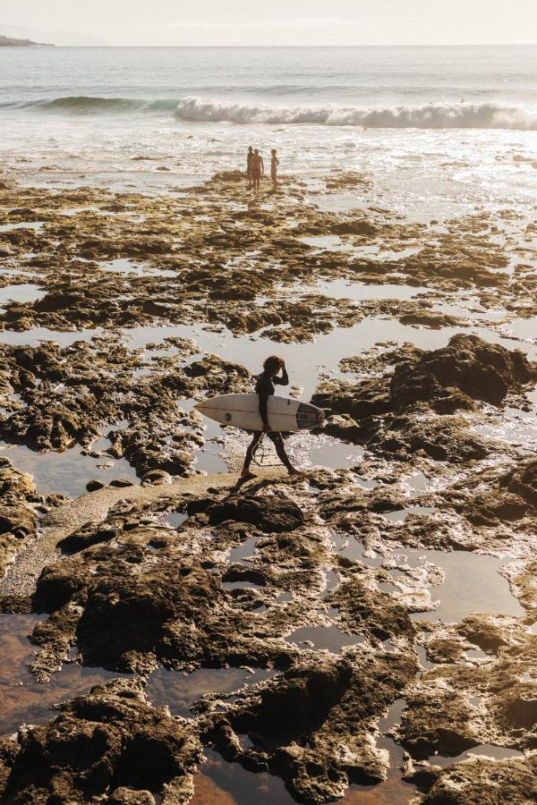 11 UNIQUE Boutique Hotels in Tenerife 11 A surfer carrying a surfboard walks across rocky tide pools toward the ocean, while a group of people stands together near the waves in the background under bright sunlight, capturing the adventurous spirit often found near boutique hotels in Tenerife.