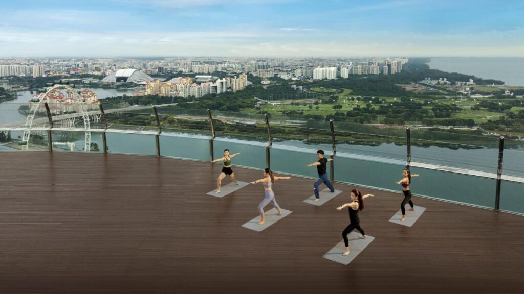 13 Most Unique Hotels in Singapore for an Incredible Holiday 6 Five people practice yoga on a rooftop deck at one of the unique hotels in Singapore, overlooking a city skyline, river, and greenery beneath a blue sky. Tall buildings and a Ferris wheel enhance the stunning urban view.