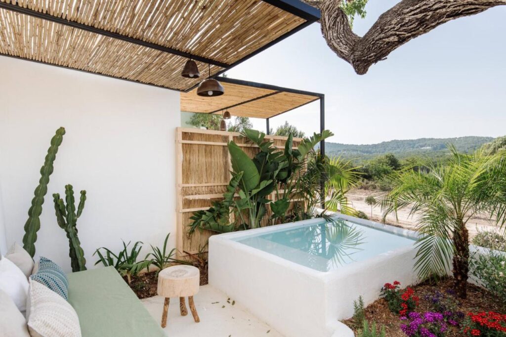 10 AUTHENTIC Finca Hotels in Ibiza 5 A small white plunge pool surrounded by tropical plants, a green cushioned bench, and a rustic wooden stool on a patio with a bamboo-covered pergola—this serene spot captures the laid-back charm of finca hotels in Ibiza, overlooking hills and trees.