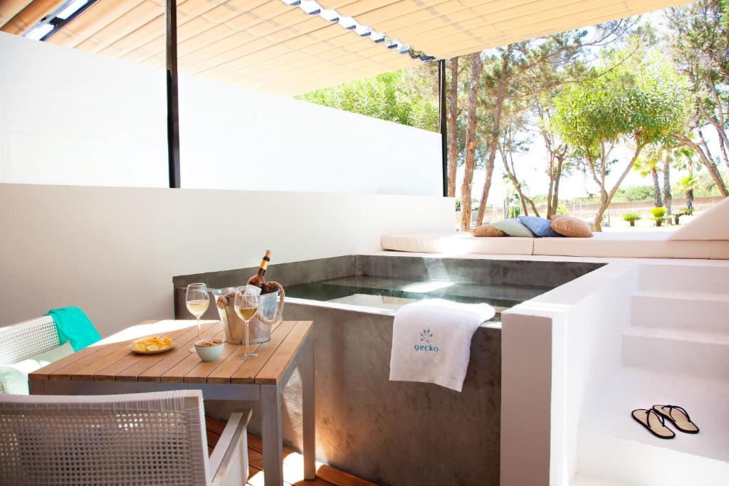 A small outdoor patio, reminiscent of boutique hotels in Formentera, features a plunge pool, wooden table with drinks and snacks, cushioned lounging area, white stairs, and trees beyond. Sunlight filters through a slatted roof.