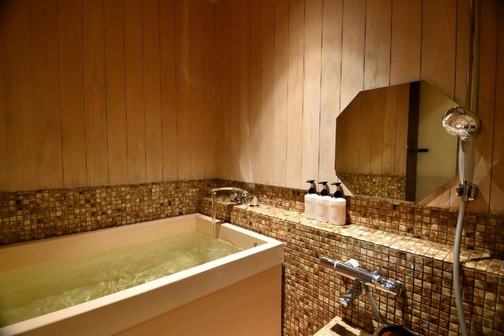7 UNIQUE Ryokans in Gion Kyoto 24 A cozy bathroom inspired by ryokans in Gion Kyoto, featuring wood-paneled walls, a deep soaking tub filled with water, mosaic tile accents, a wall-mounted showerhead, and toiletries on the tiled ledge beside a polygonal mirror.