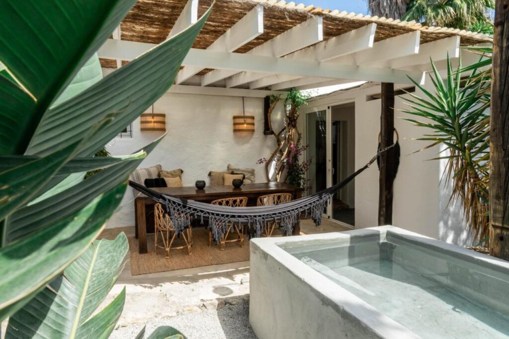10 AUTHENTIC Finca Hotels in Ibiza 8 A cozy patio reminiscent of finca hotels in Ibiza features a hammock, a wooden dining table with chairs, a small concrete pool, potted plants, and a shaded pergola with woven roofing for a relaxed, inviting outdoor space.