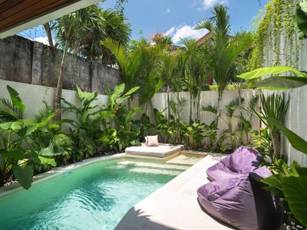 14 UNIQUE Boutique Hotels in Canggu, Bali 55 A small, private backyard pool surrounded by lush tropical plants and palm trees, with a built-in sunbed, a cushion, and two purple bean bag chairs on the stone patio—echoing the tranquil vibes of boutique hotels in Canggu. The sky is clear and sunny.