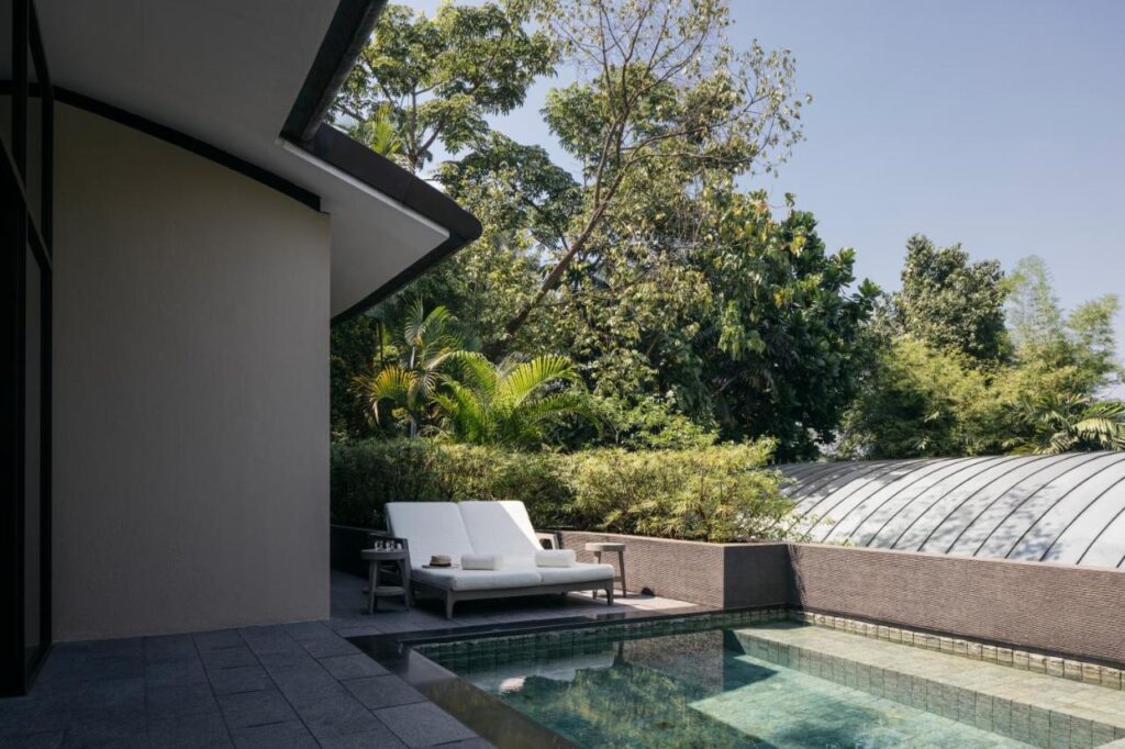 13 Most Unique Hotels in Singapore for an Incredible Holiday 53 A modern outdoor patio at one of the unique hotels in Singapore features a lounge chair and table beside a clear swimming pool, surrounded by lush greenery and tall trees under a clear blue sky.